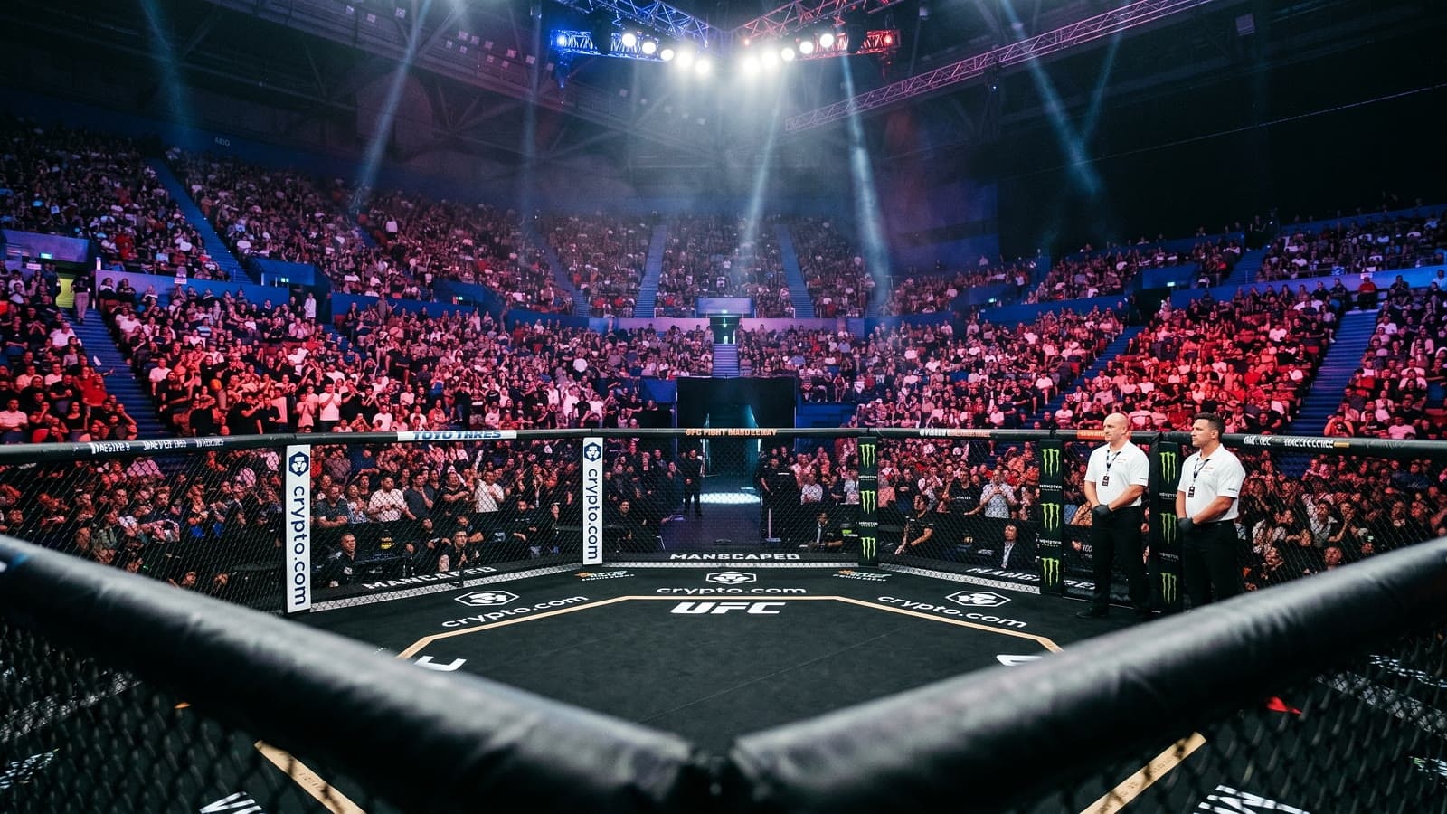 Cage-side view of the UFC octagon inside RAC Arena, Perth, Western Australia, with two referees at the cage edge and a packed crowd in the stadium bowl beyond.